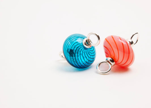 Close up front view of a pair blown glass bead charm left to right: cyan with blue swirls and pink with red swirls both with Sterling Silver looped endings on a white background.