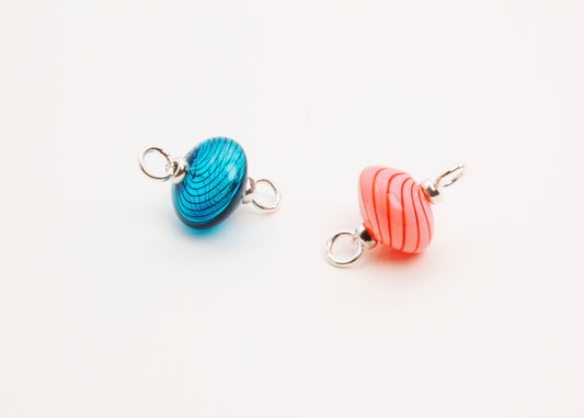 Close up front view of a pair blown glass bead charm left to right: cyan with blue swirls and pink with red swirls both with Sterling Silver looped endings placed diagonally on a white background.