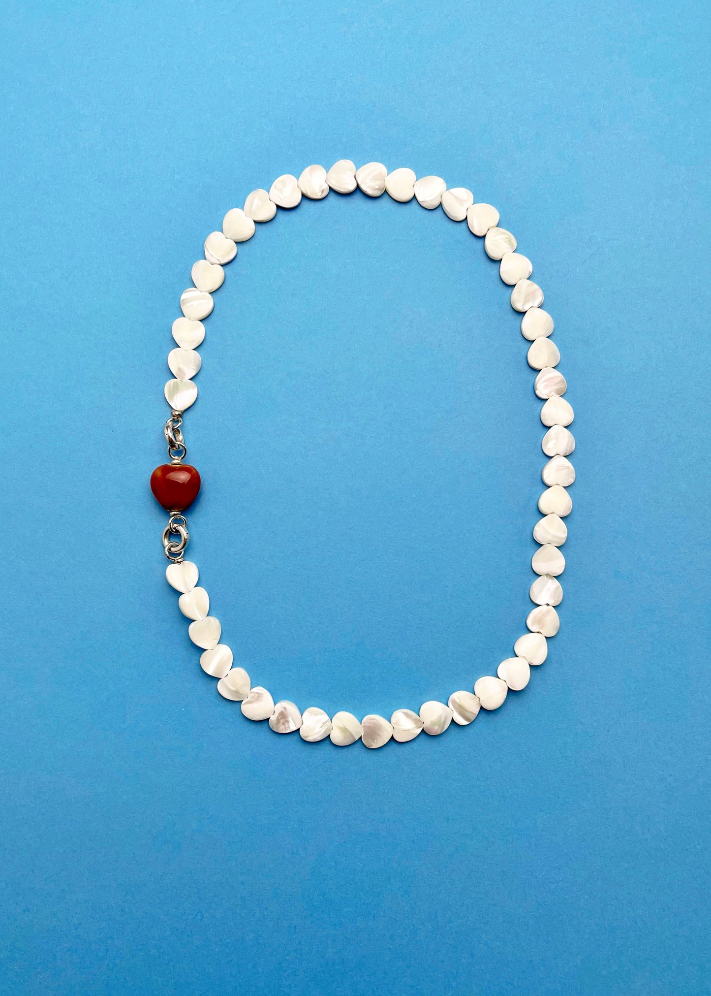Overhead view of a Mother of Pearl heart beads necklace with a red chunky porcelain heart bead charm and Sterling Silver endings placed on a cyan background