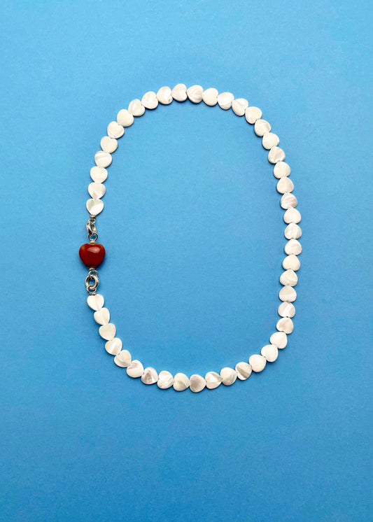 Overhead view of a Mother of Pearl heart beads necklace with a red chunky porcelain heart bead charm and Sterling Silver endings placed on a cyan background