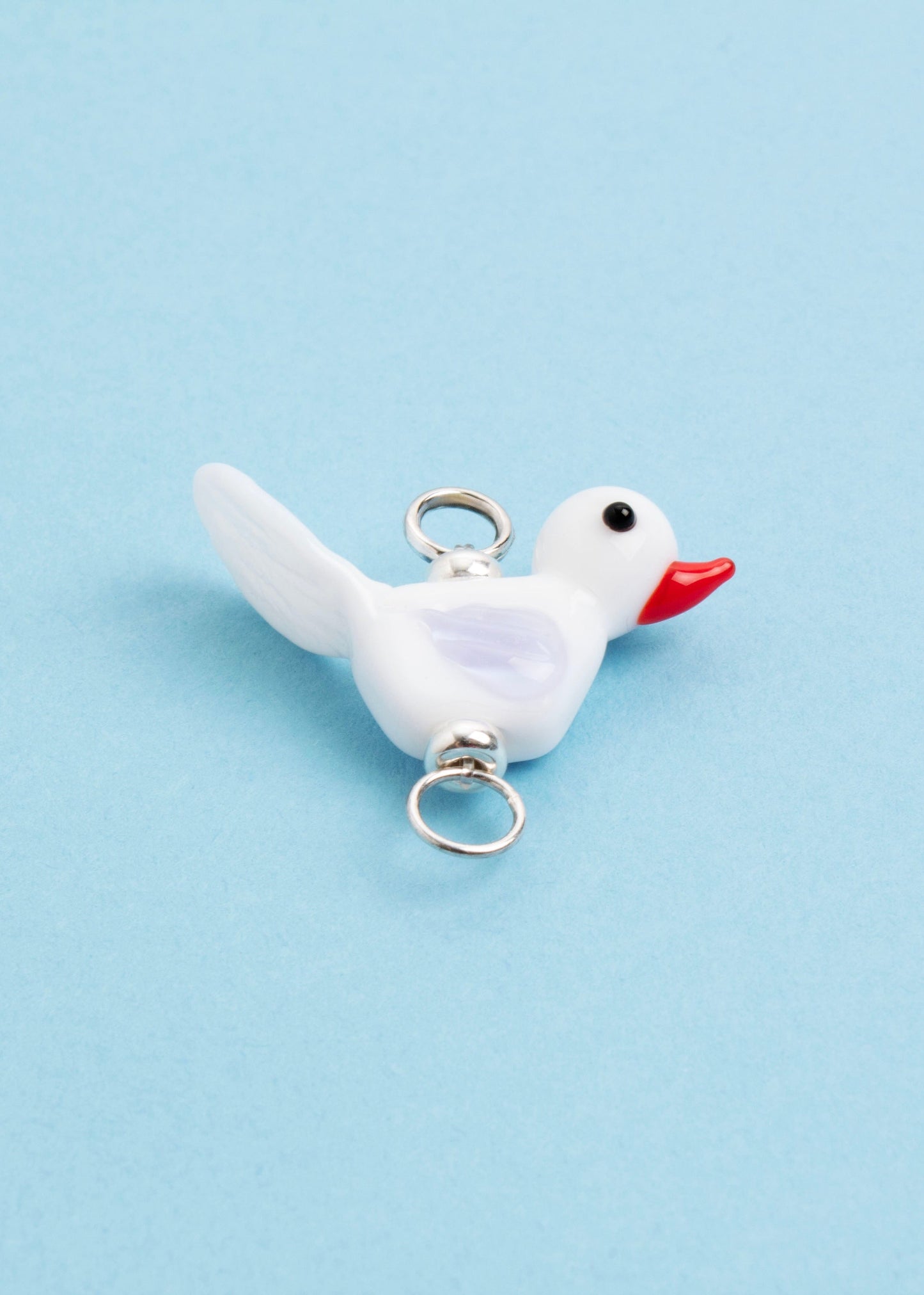 Front view of a white_lamp_work_bird charm_with an orange beak and lavender wings on a sky blue background