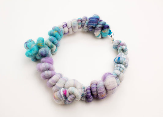 Plan view of a closed wool yarn necklace in cream, turquoise, purple and hazelnut accents with Sterling Silver looped endings on a white background