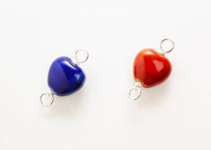 Front close up of two glazed porcelain heart charm in blue and red form left to right, both with sterling silver looped endings on a white background. 