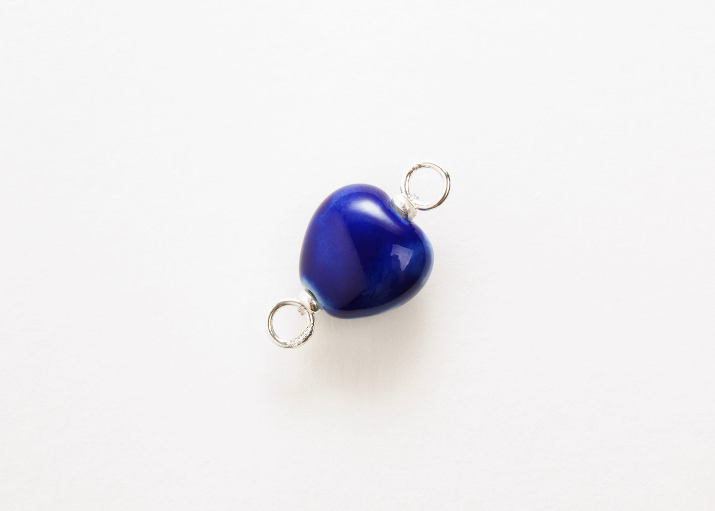 Diagonal close up of a blue glazed porcelain heart-shaped charm  with sterling silver looped endings on a white background. 