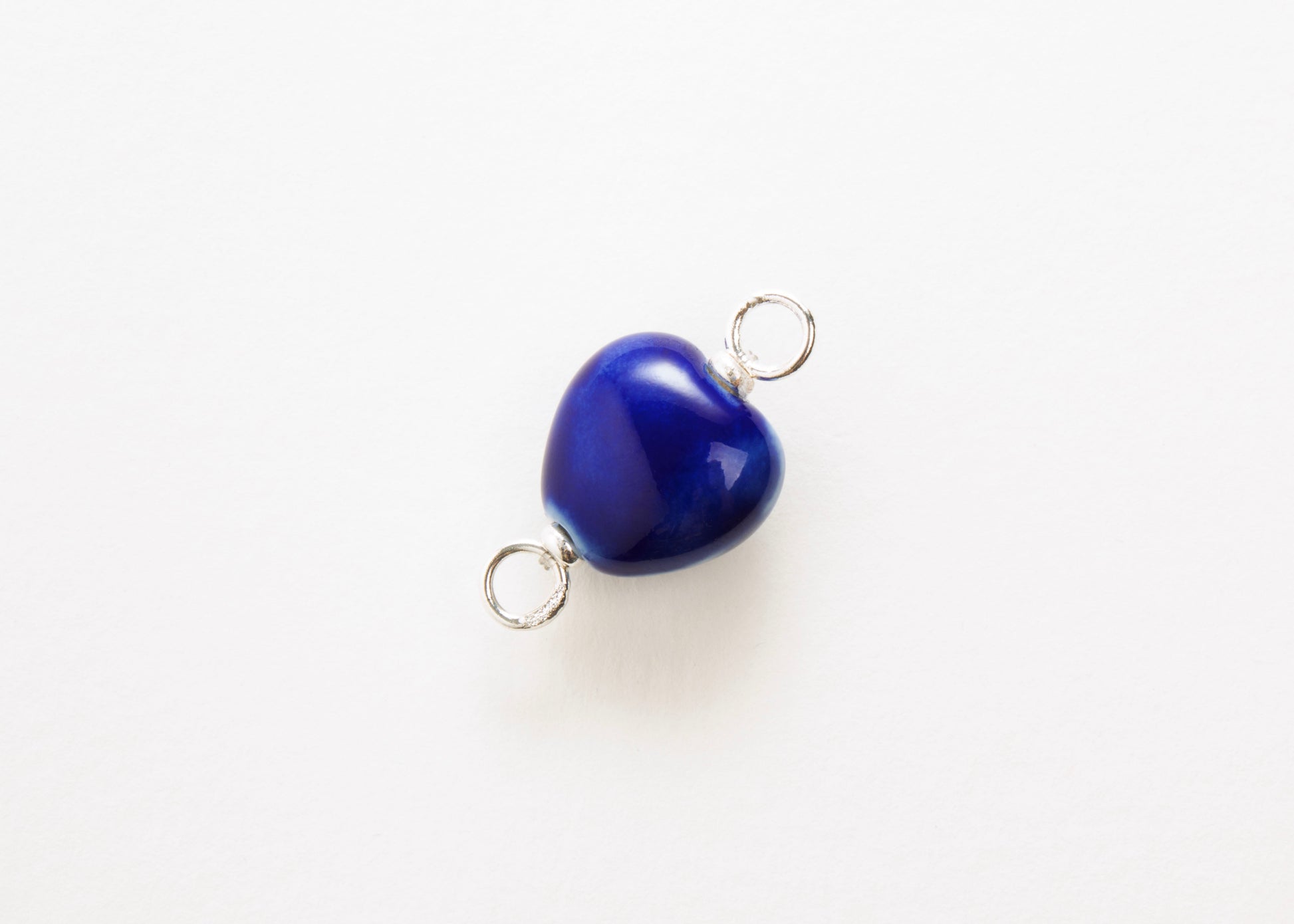 Diagonal close up of a blue glazed porcelain heart-shaped charm  with sterling silver looped endings on a white background. 