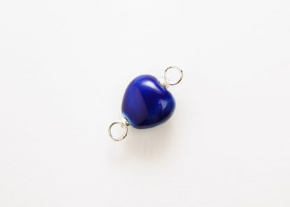 Diagonal close up of a blue glazed porcelain heart-shaped charm  with sterling silver looped endings on a white background. 
