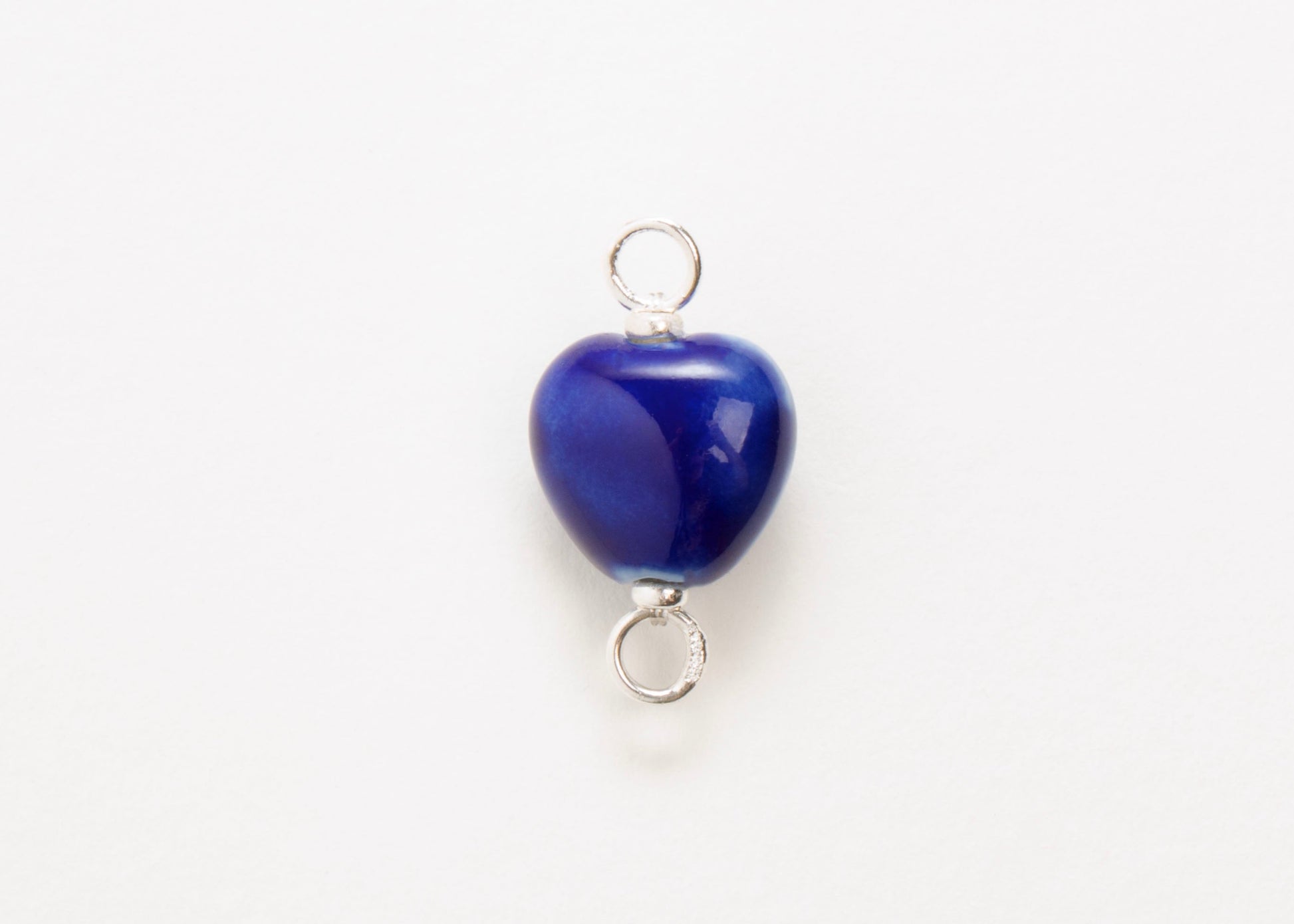 Front close up of a blue glazed porcelain heart-shaped charm  with sterling silver looped endings on a white background. 