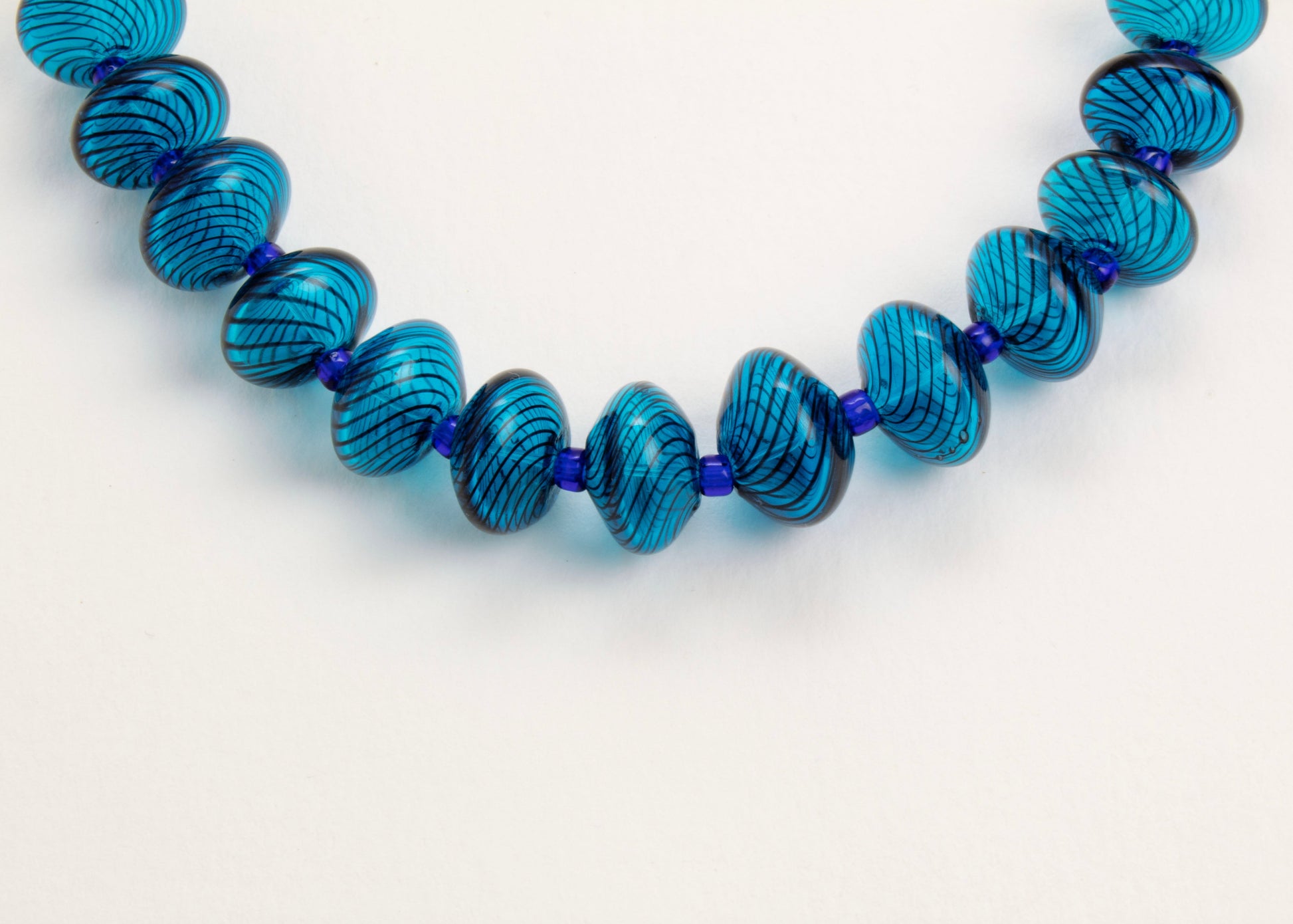 Close up of the drop detail of a cyan blown glass bead strand with dark blue  swirls and electric blue separators on white background