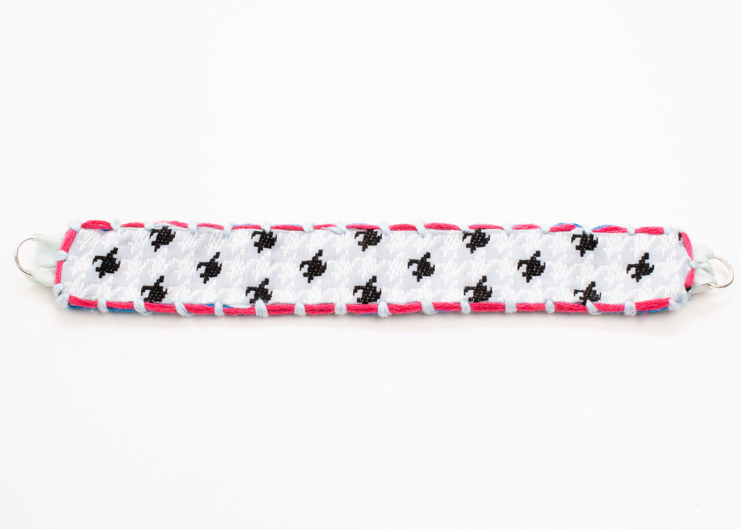 Plan view of a reversible cotton woven strap bracelet in houndstooth pattern in grey blue with embroidered borders in  fuchsia and light blue, and Sterling Silver looped endings detail on a white background.