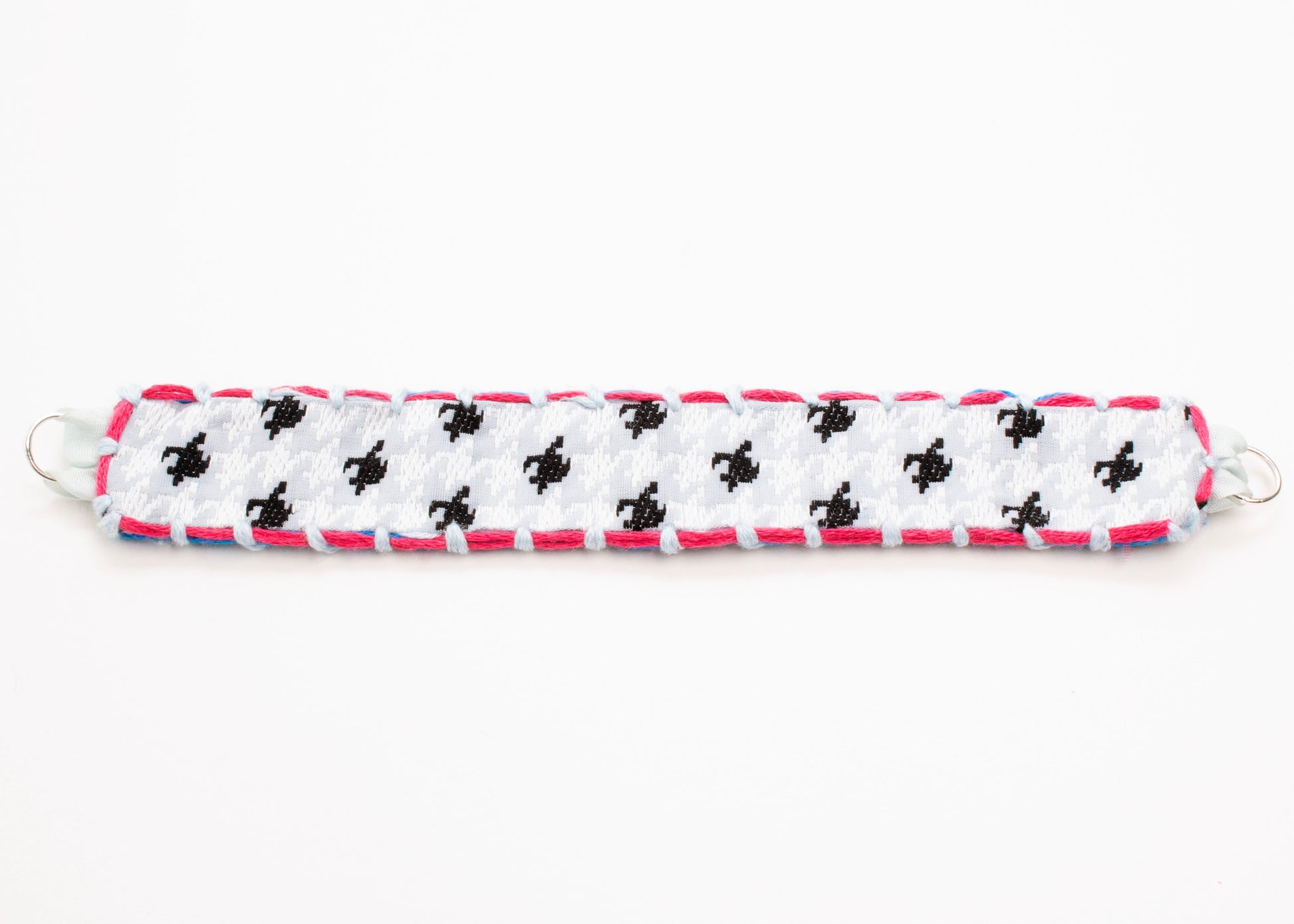 Plan view of a reversible cotton woven strap bracelet in houndstooth pattern in grey blue with embroidered borders in  fuchsia and light blue, and Sterling Silver looped endings detail on a white background.