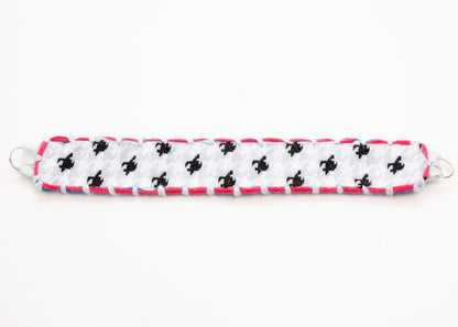 Plan view of a reversible cotton woven strap bracelet in houndstooth pattern in grey blue with embroidered borders in  fuchsia and light blue, and Sterling Silver looped endings detail on a white background.