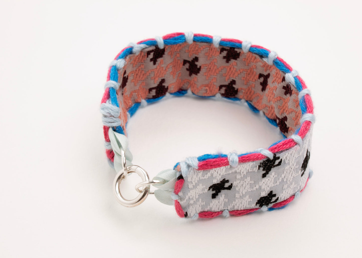 Front view of a reversible cotton woven strap bracelet in houndstooth pattern in terracotta and silver blue with embroidered borders in cyan and fuchsia, and Sterling Silver looped endings detail on a white background.
