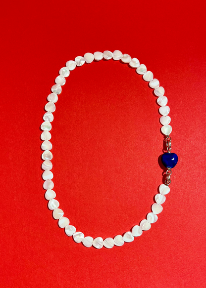 Overhead view of an MOP heart carved  beaded necklace with a chunky blue porcelain charm attached both with Sterling Silver endings on a solid red background.