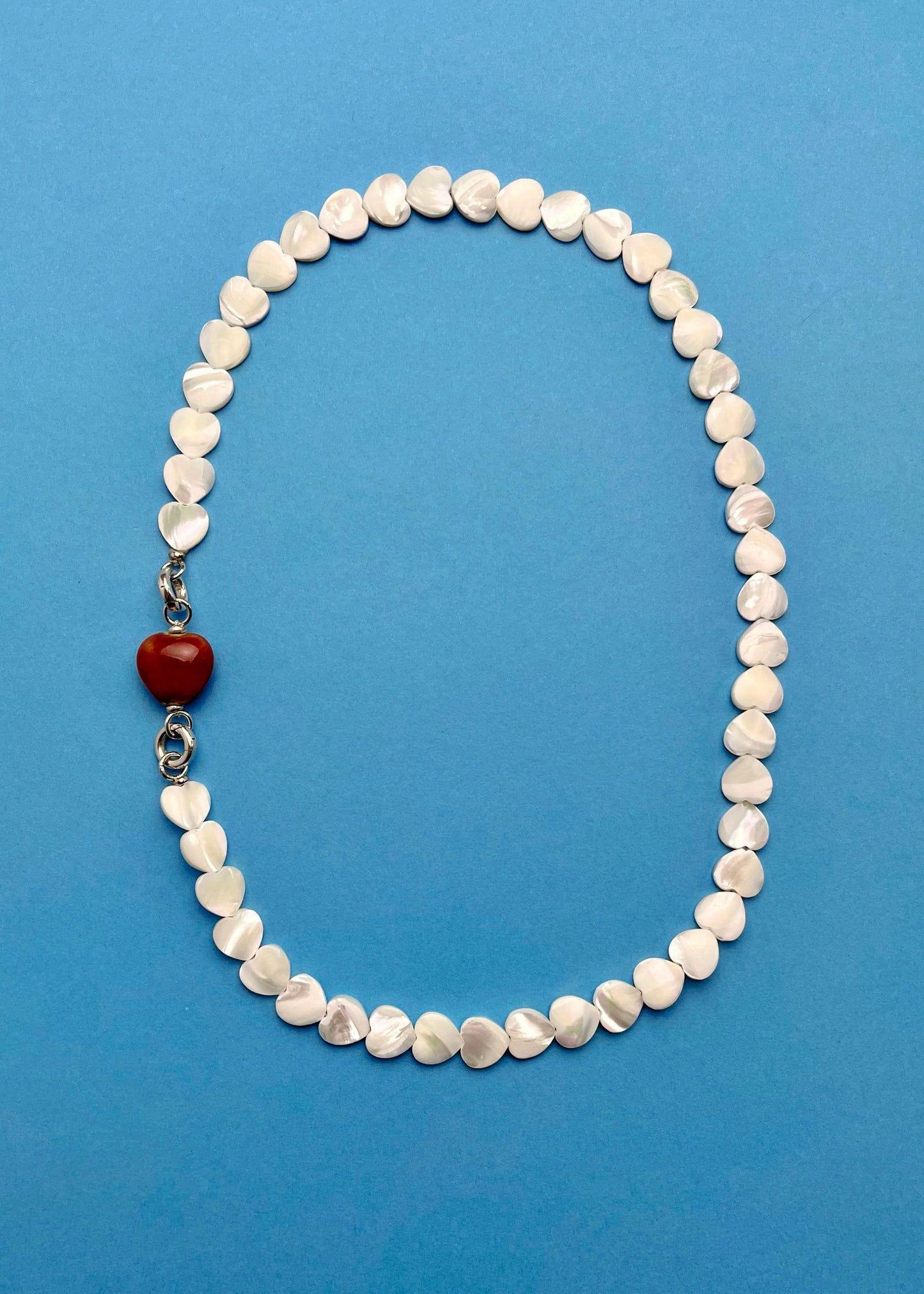 Overhead view of an MOP_necklace_with_red_porcelain_heart_charm attached and Sterling Silver endings _placed on a cerulean background