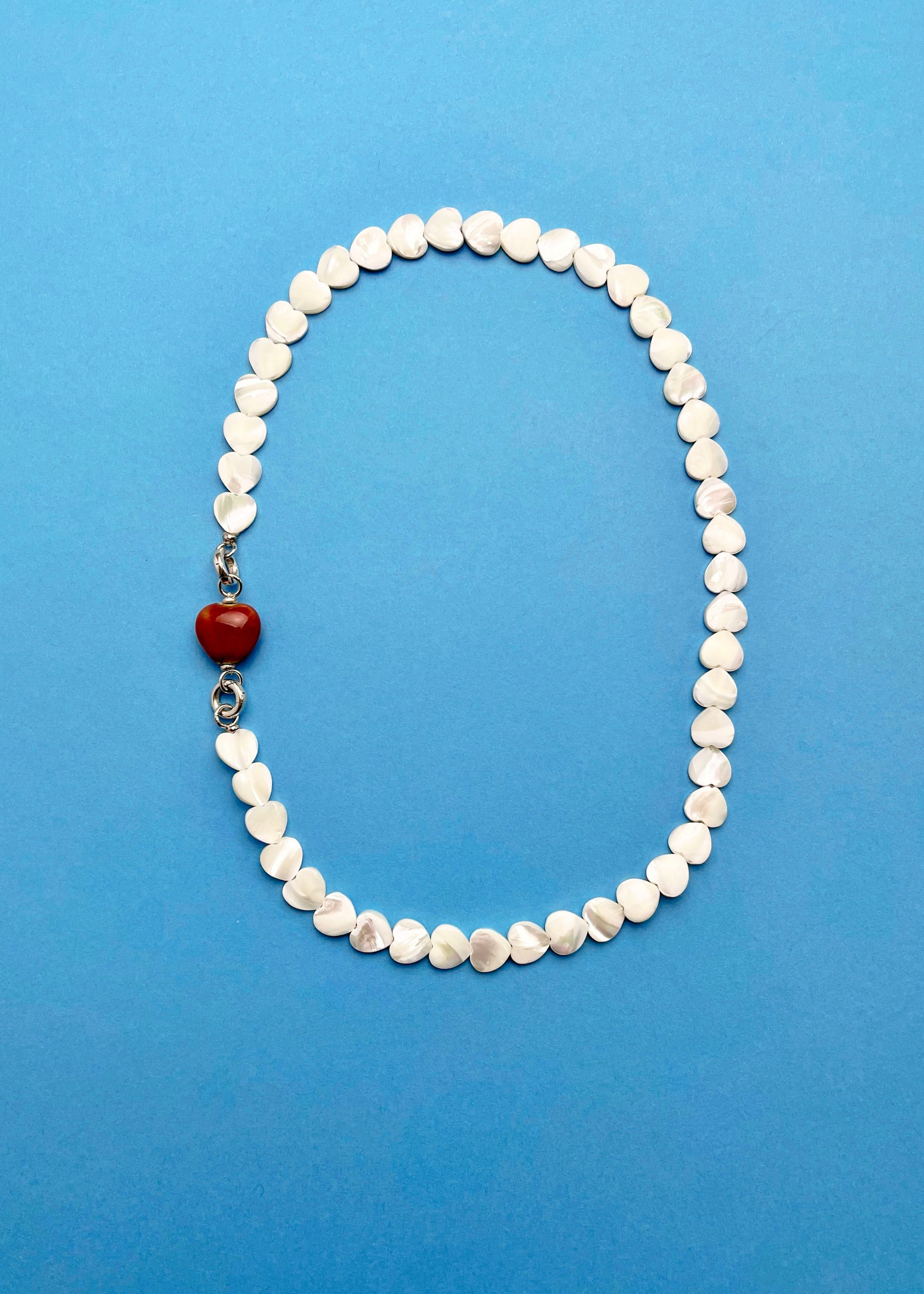 Overhead view of a Mother of Pearl heart beads necklace with a red chunky porcelain heart bead charm and Sterling Silver endings placed on a cyan background
