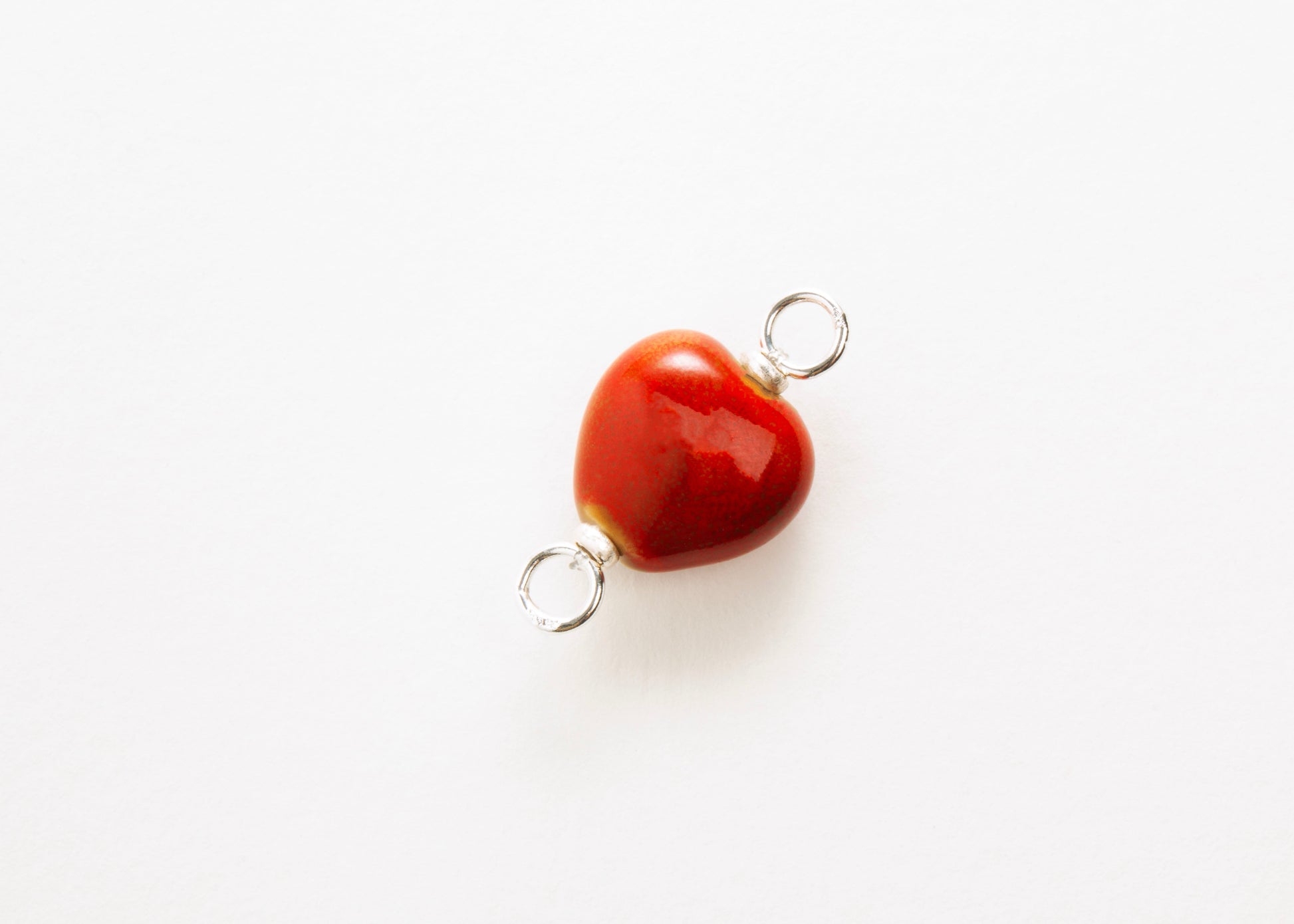 Diagonal close up of a red glazed porcelain heart-shaped charm with sterling silver looped endings on a white background. 