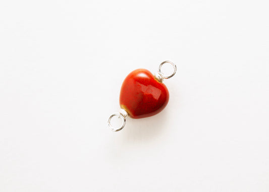 Diagonal close up of a red glazed porcelain heart-shaped charm with sterling silver looped endings on a white background. 