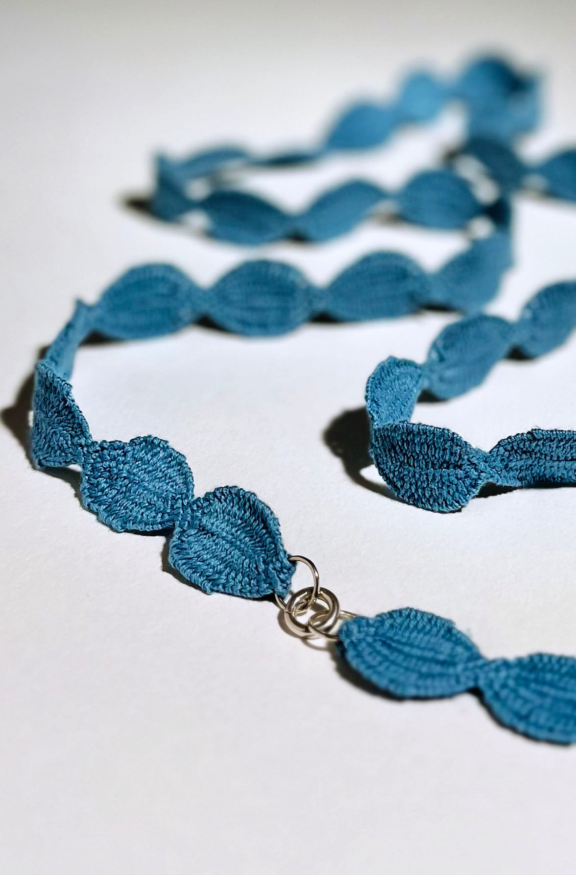 Close view of the Sterling Silver endings of an interwoven cotton ovals necklace in sky blue laid on a white background.