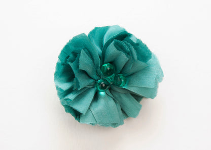Front view of a teal silk flower charm with glass detail in center on a white background.