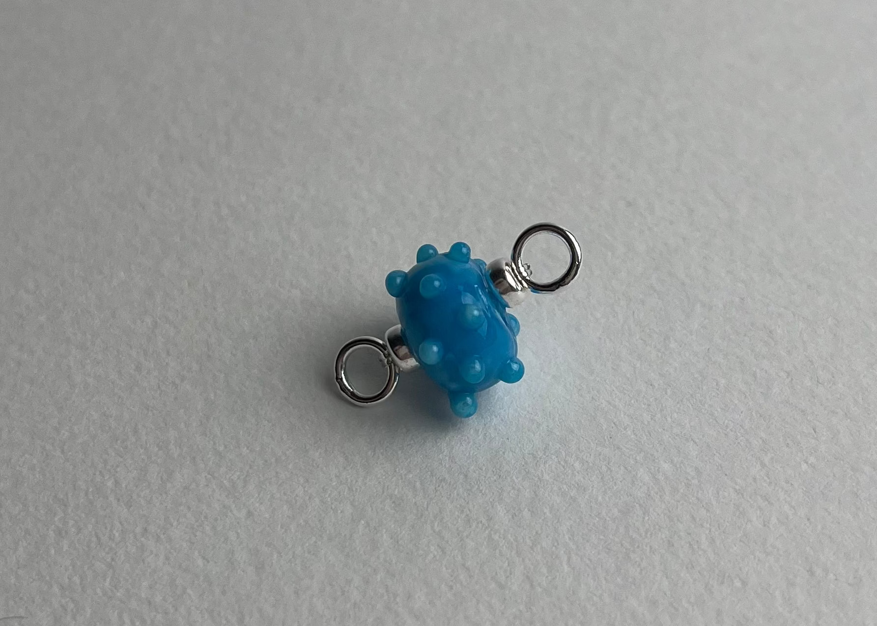 Close up of a blue urchin shaped bumpy lampwork charm with sterling silver ends on a white background