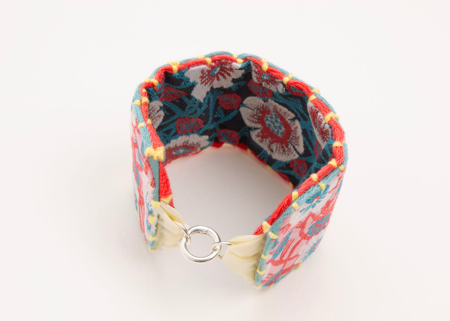 Front view of a closed cotton woven reversible strap bracelet, with flower motifs and embroidered borders in persimmon and pink, teal and deep blue with Sterling silver looped endings on a white background.