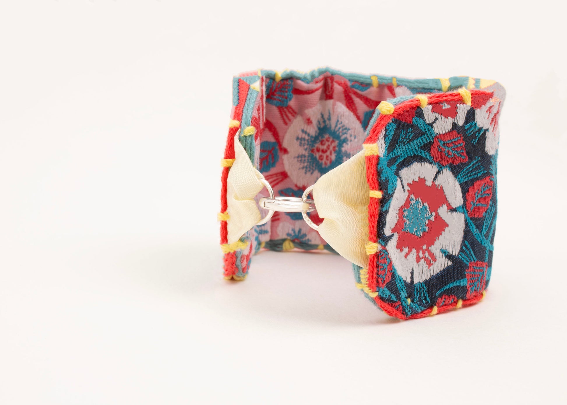 Front view of a cotton woven reversible strap bracelet, with flower motifs and embroidered borders in persimmon and pink, teal, deep blue and yellow with Sterling silver looped endings on a white background.