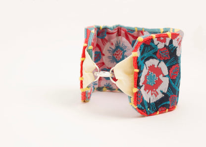 Front view of a cotton woven reversible strap bracelet, with flower motifs and embroidered borders in persimmon and pink, teal, deep blue and yellow with Sterling silver looped endings on a white background.