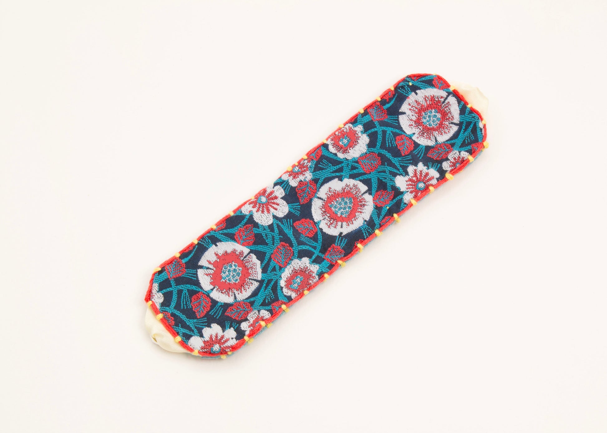 Plan view of a cotton woven reversible strap bracelet, with flower motifs and embroidered borders in persimmon and pink, teal, deep blue and yellow with Sterling silver looped endings on a white background.