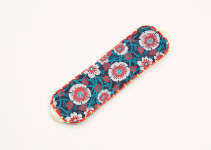 Plan view of a cotton woven reversible strap bracelet, with flower motifs and embroidered borders in persimmon and pink, teal, deep blue and yellow with Sterling silver looped endings on a white background.