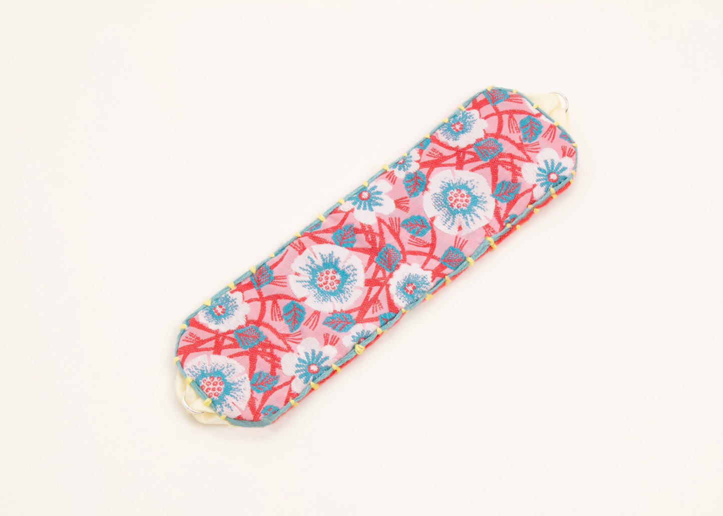 Plan view of a cotton woven reversible strap bracelet, with flower motifs and embroidered borders in persimmon and pink, teal and butter yellow with Sterling silver looped endings on a white background.
