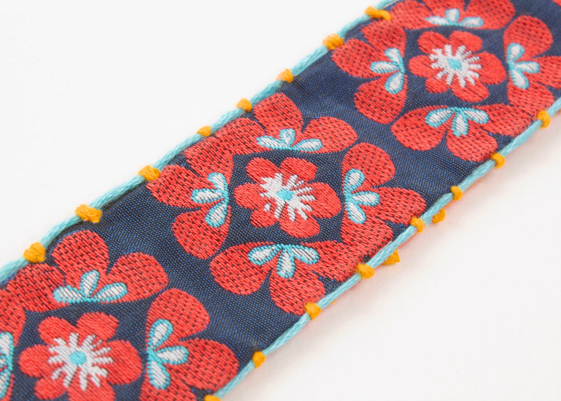 Close up of fabric detail of a cotton woven reversible strap bracelet, with squarish flower motifs in teal, deep blue and mellow yellow with Sterling silver looped endings on a white background.