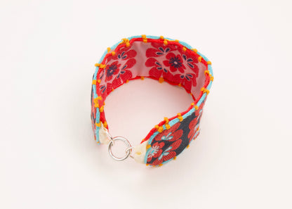 Plan view of a closed cotton woven reversible strap bracelet, with squarish flower motifs in persimmon, pink, teal deep blue and mellow yellow with Sterling silver looped endings on a white background.