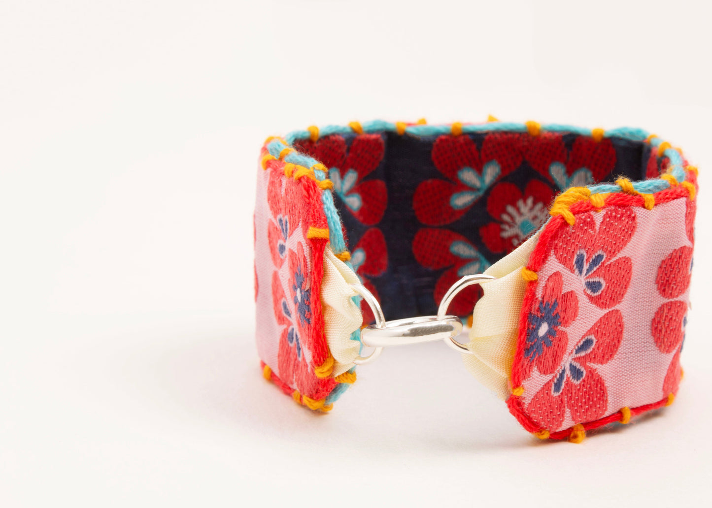 Front view of a cotton woven reversible strap bracelet, with squarish flower motifs  in persimmon, pink, teal ,deep blue and mellow yellow with Sterling silver looped endings on a white background.