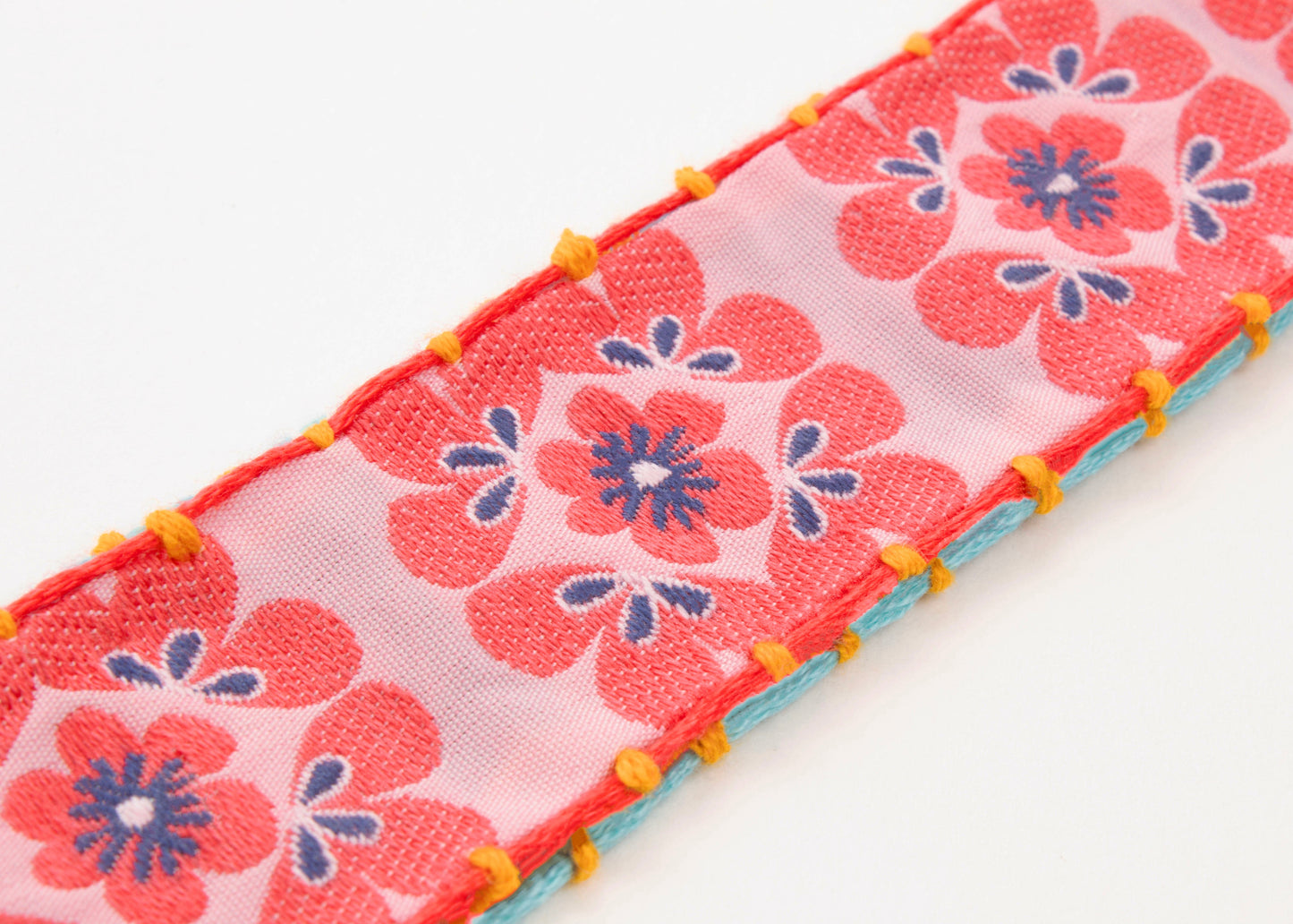 Close up of fabric detail of a cotton woven reversible strap bracelet, with squarish flower motifs in persimmon and pink and mellow yellow with Sterling silver looped endings on a white background.