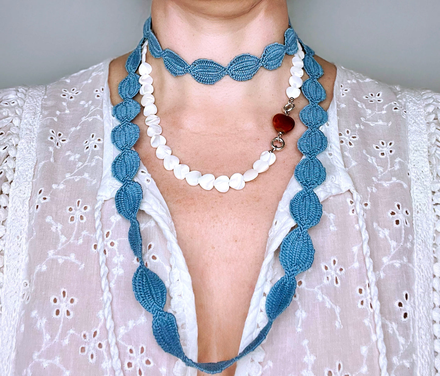 Front view of a woman wearing an interwoven cotton ovals necklace in sky blue double looped in choker style layered with a heart shape MOP bead necklace with porcelain red chunky heart charm