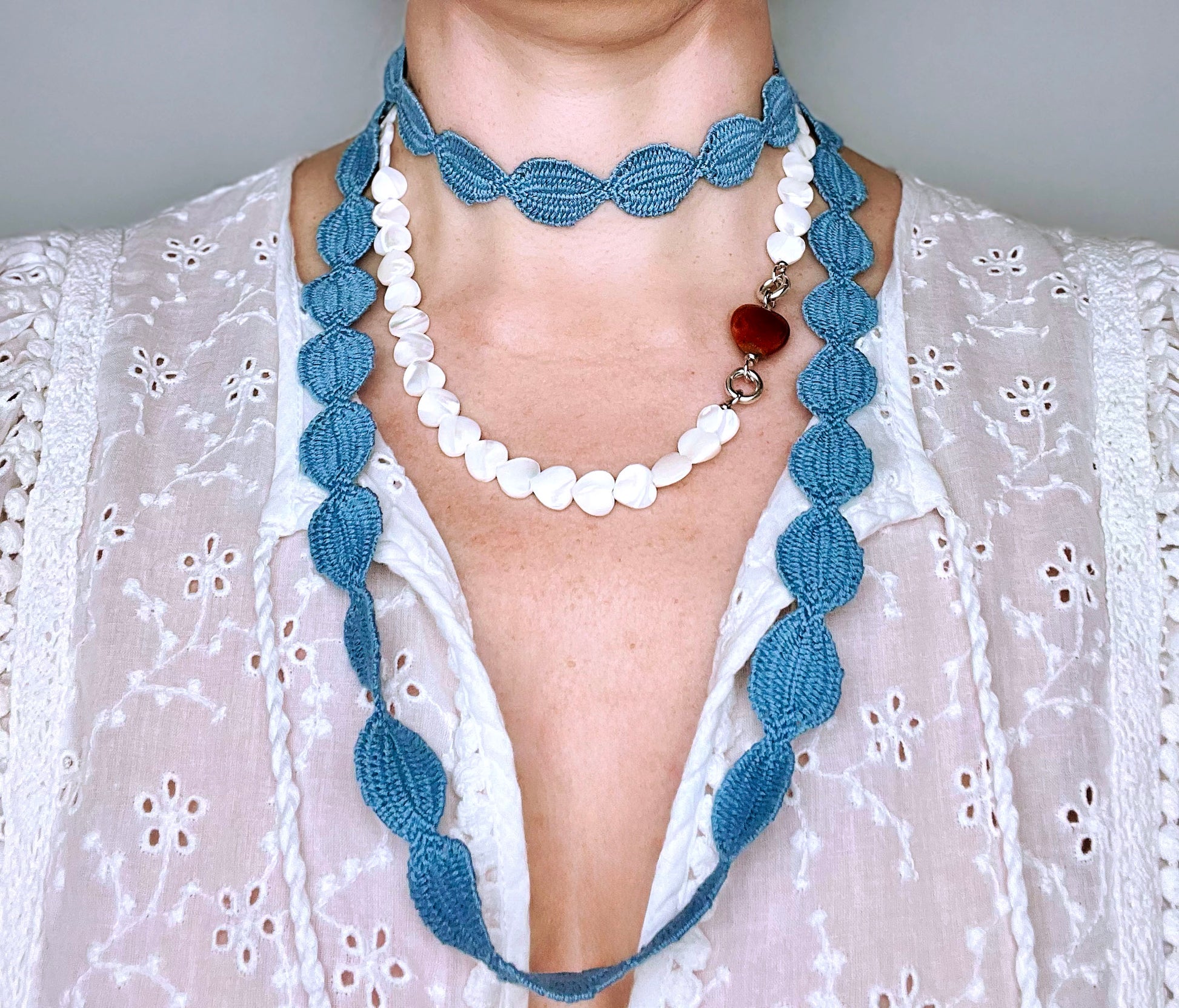 Front view of a woman wearing an interwoven cotton ovals necklace in sky blue double looped in choker style layered with a heart shape MOP bead necklace with porcelain red chunky heart charm