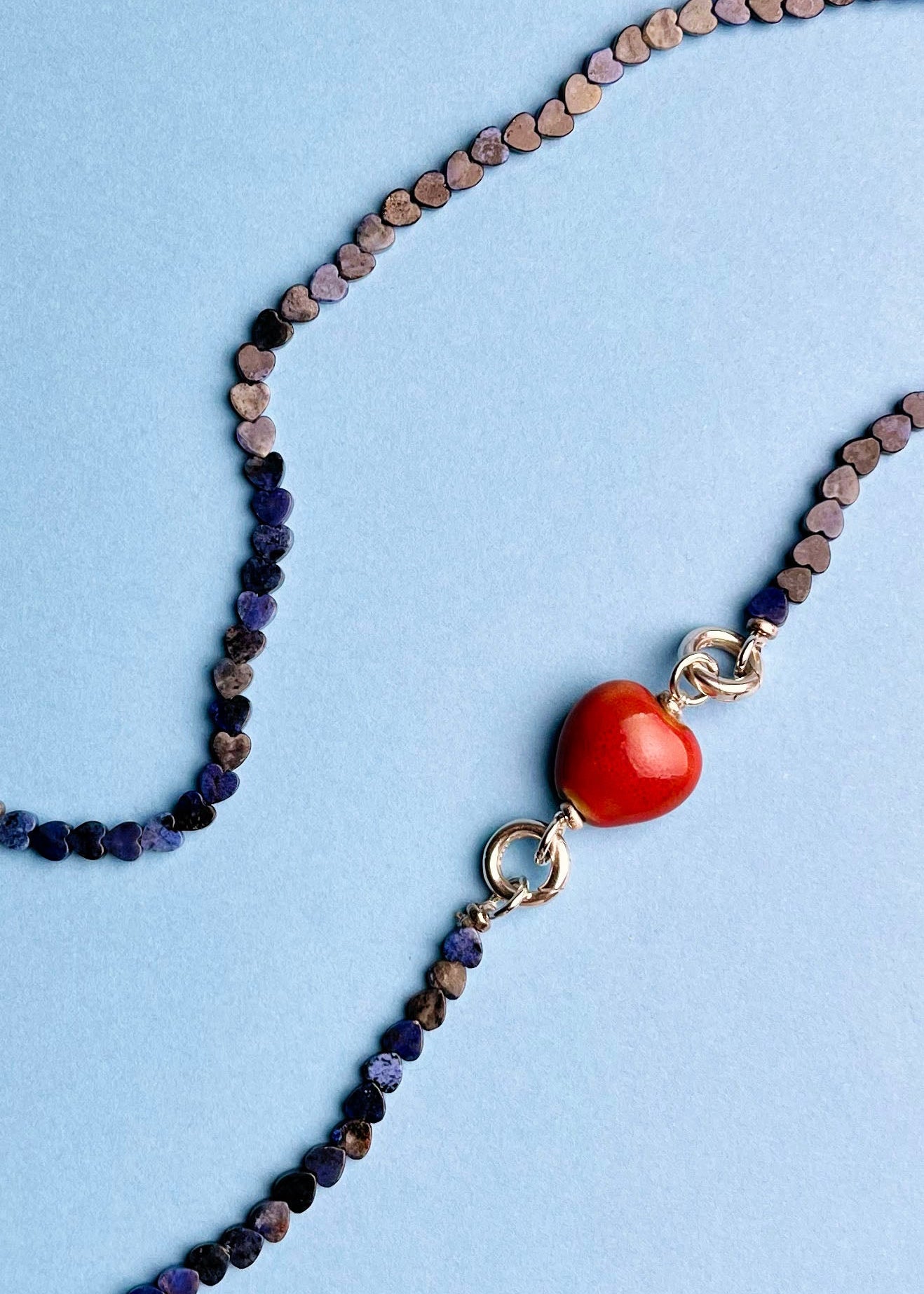 Overhead detail of a red chunky porcelain charm attached to a lapis lazuli heart shaped carved beads necklace with Sterling Silver endings placed in two loops on a navy blue background