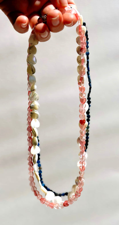Hand holding a stack of gemstone heart carved beads, in cherry quartz, MOP and laid lazuli, al with Sterling Silver endings on a white background