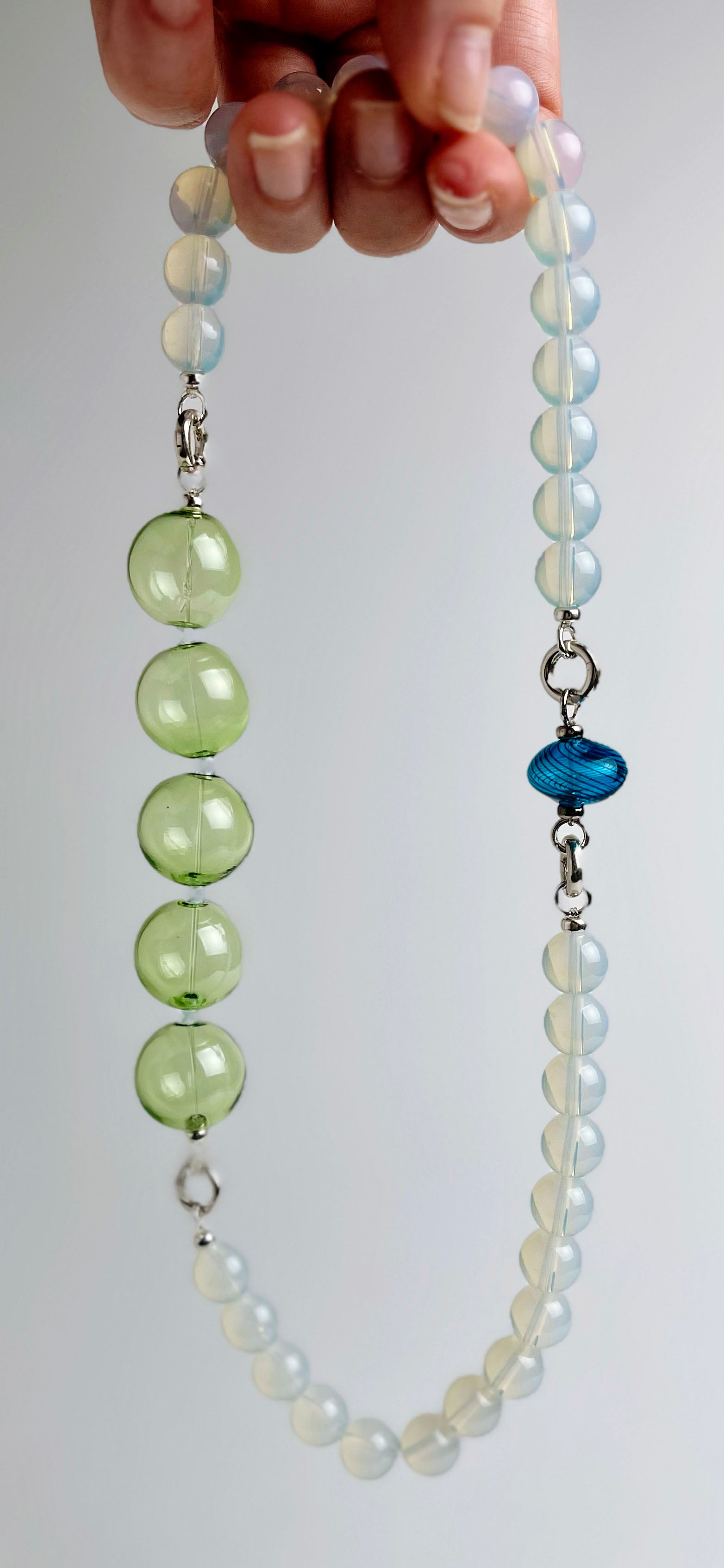 Front view of a hand holding a ready to wear necklace made with green blue Opalite chainlets, green chunky glass bead drop and a cerulean bumpy glass charm with Sterling Silver endings on a light grey  backdrop