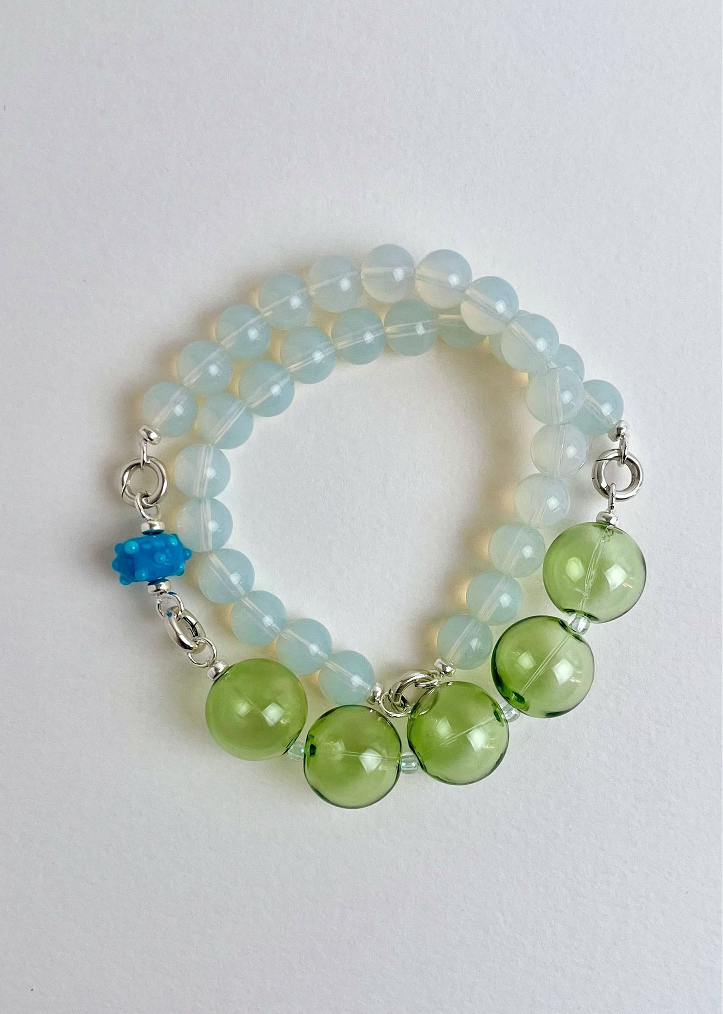 Overhead view of a ready to wear necklace made with green blue Opalite chainlets, green chunky glass bead drop and a cerulean bumpy glass charm with Sterling Silver endings on a white background