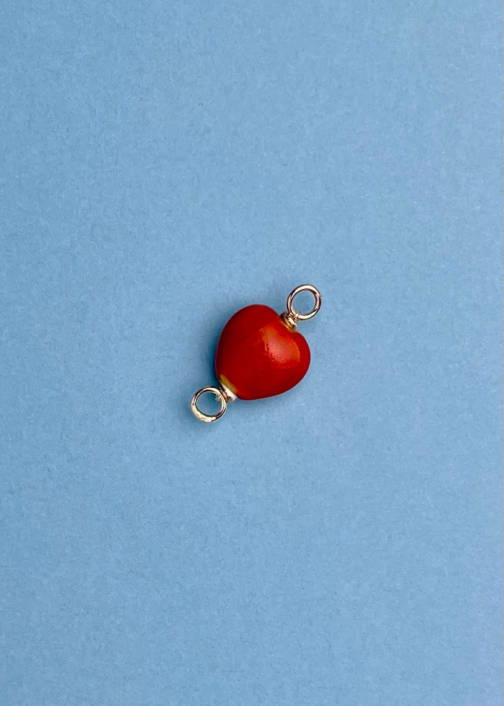 Overhead view of a red_chunky_porcelain_heart charm with Sterling Silver endings placed _diagonally on a light blue background