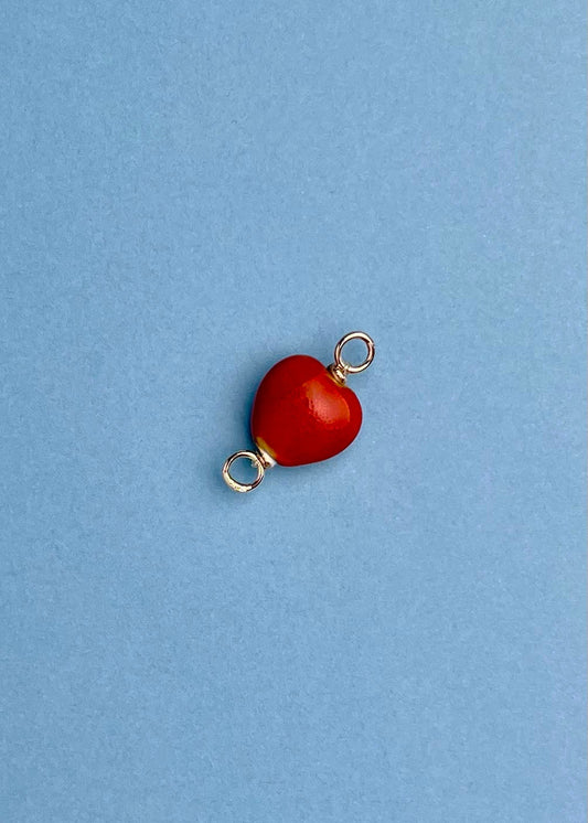 Overhead view of a red_chunky_porcelain_heart charm with Sterling Silver endings placed _diagonally on a light blue background