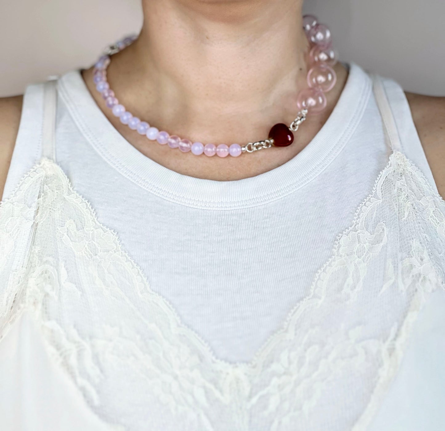 Front view of a ready to wear necklace on a round neck line made with peach pink opaline chainlets,  a pink chunky glass bead drop and red heart charm with Sterling Silver endings