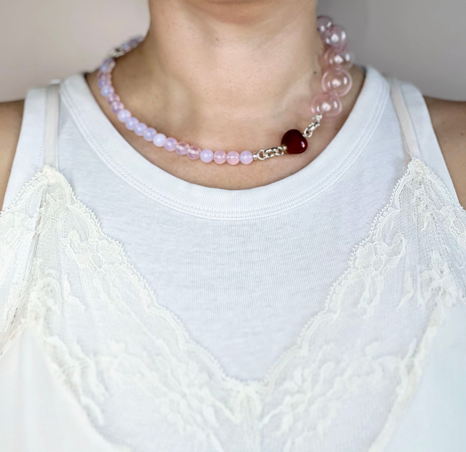 Front view of a ready to wear necklace on a round neck line made with peach pink opaline chainlets,  a pink chunky glass bead drop and red heart charm with Sterling Silver endings