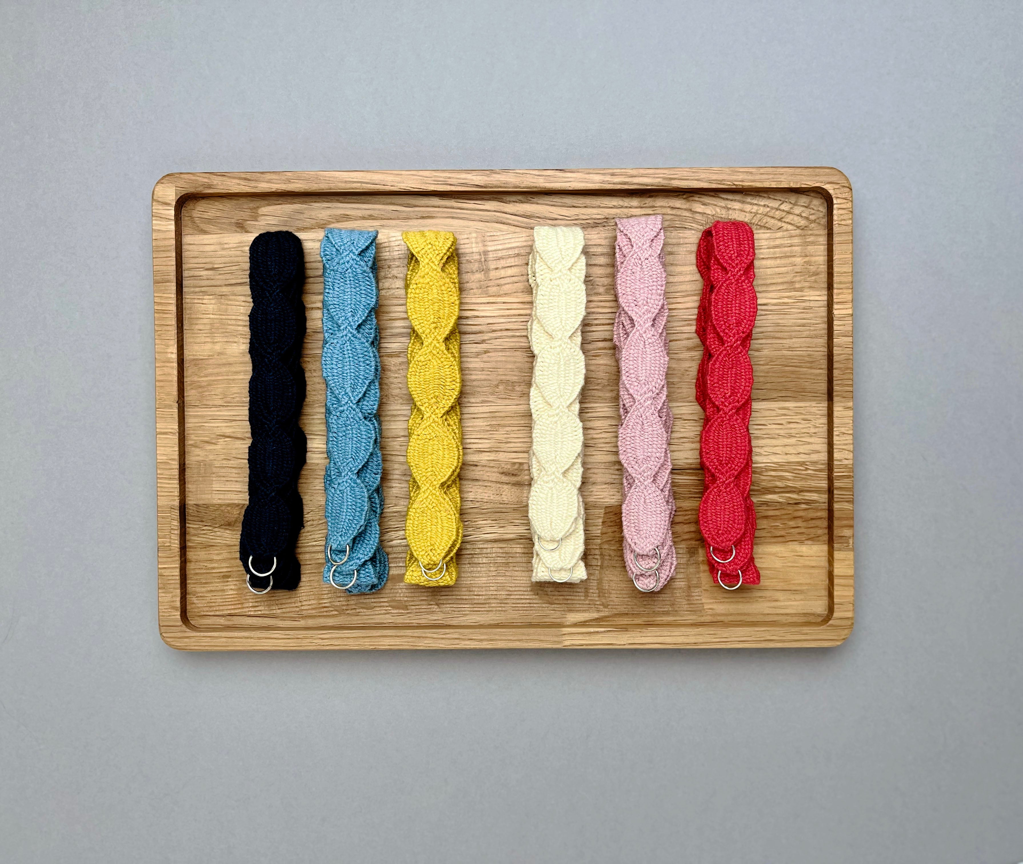 Overhead view of six cotton necklaces with interwoven ovals in navy, soft blue, yellow, cream, pink, and red arranged vertically on a rectangular wooden tray against a solid gray background.