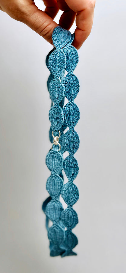 Side handheld view of a sky blue interwoven cotton ovals necklace in a double loop on a grey background