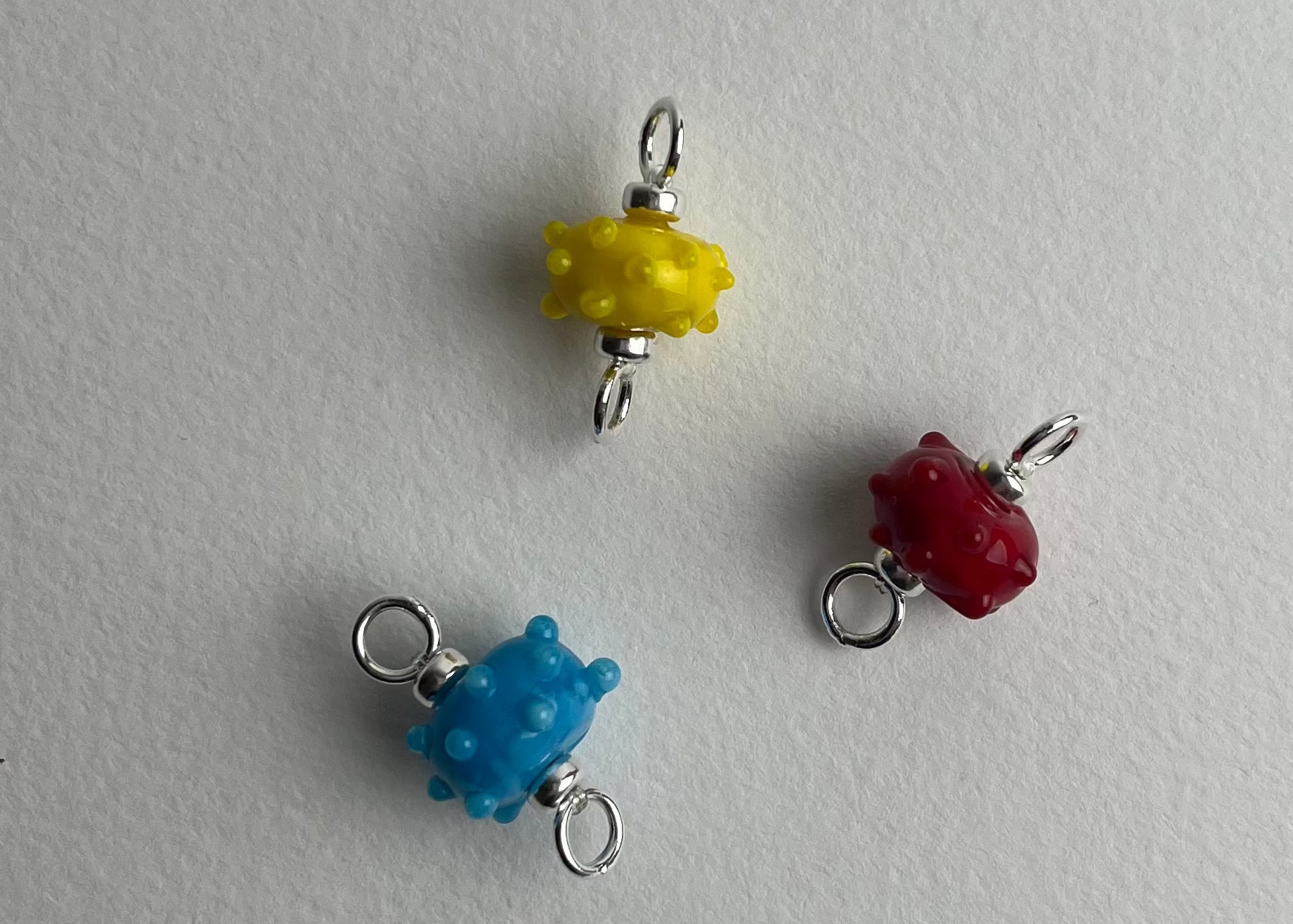 Close up of trio of urchin shaped bumpy lampwork charms with sterling silver ends in blue, red and yellow on a white background