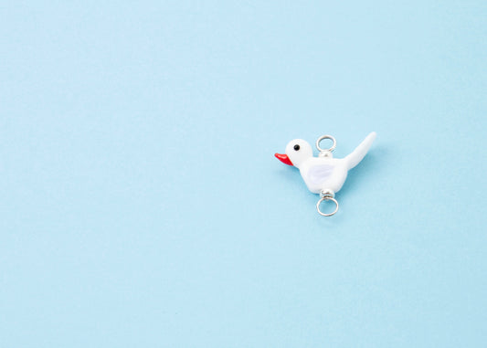 Plan view left side of a white_lampwork_bird charm_with an orange beak and lavender wings on a sky blue background