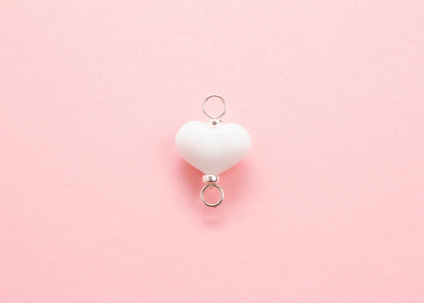 Plan view of white lampwork glass heart charm with Sterling Silver looped endings on a pink background