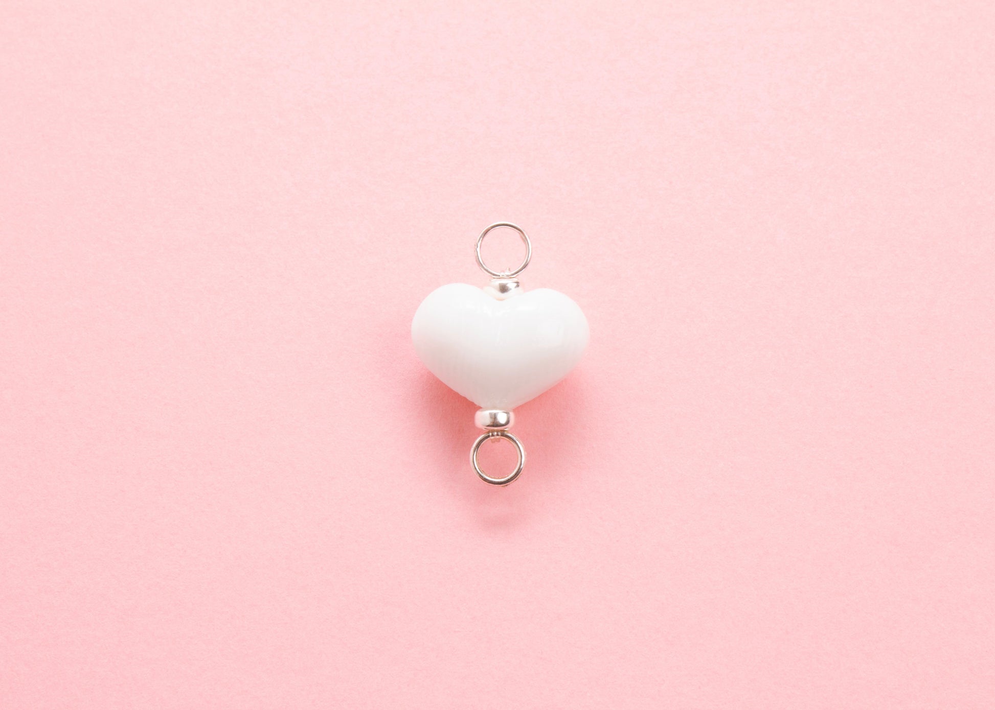 Plan view of white lampwork glass heart charm with Sterling Silver looped endings on a pink background
